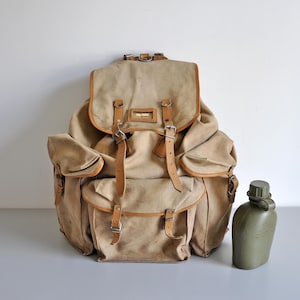 Pode incluir: Uma mochila de lona bege com acabamento em couro, vários bolsos externos com fechos de fivela e uma aba superior. Ao lado da mochila, há um cantil de plástico verde-oliva com tampa de rosca e texto moldado na superfície frontal.