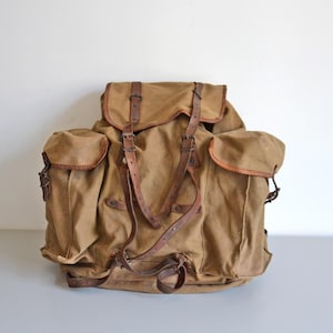 Peut inclure: Un sac à dos en toile beige doté d'un grand compartiment principal avec un rabat, deux poches latérales à rabat et des lanières en cuir marron avec des boucles en métal. Le design comprend des bretelles réglables en cuir et des ferrures métalliques.