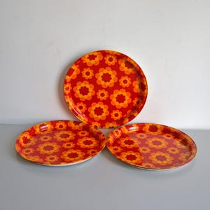 Op de afbeelding: Drie ronde metalen dienbladen met een levendige rode achtergrond en een herhalend patroon van oranje en gele gestileerde bloemen. De dienbladen hebben een gladde, glanzende afwerking en opstaande randen, gerangschikt met één dienblad achter twee andere.