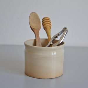Peut inclure: Un pot de cuisine en céramique beige contenant une cuillère en bois, un bâtonnet à miel et un casse-noix argenté. Le pot est cylindrique et repose sur une surface gris clair.