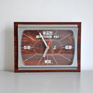 May include: A rectangular wall clock with a dark brown wood grain face and a silver metallic frame. The clock features white numerals at 3, 6, 9, and 12 o'clock, with a day and date display showing SAMEDI 25. It has silver hour and minute hands.