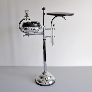 Può includere: Un posacenere da terra cromato con un'asta centrale e una base circolare. Include un posacenere nero a forma di ciotola su un lato e un piccolo tavolino nero circolare e piatto sul lato opposto, sostenuti da bracci metallici curvi.