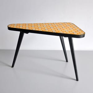 Puede incluir: Una mesa auxiliar triangular con un patrón geométrico de azulejos amarillos y blancos en la superficie superior. La mesa se apoya en tres patas negras finas y cónicas, y tiene un borde negro que rodea la superficie superior estampada.
