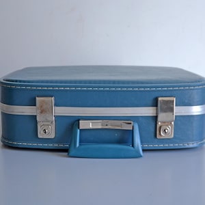 May include: A rectangular blue suitcase with a textured finish, featuring a central blue handle, a silver-toned metal trim, and two silver-toned metal locking latches on the front. The suitcase has white stitching along the top and bottom edges.