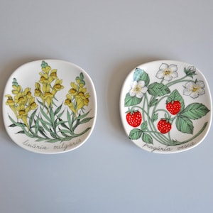 Puede incluir: Dos platos cuadrados de cerámica con diseños florales. Uno presenta flores amarillas con hojas verdes y el texto "linaria vulgaris". El otro muestra fresas, flores blancas, hojas verdes y el texto "fragaria vesca".