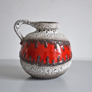 Puede incluir: Un jarrón de cerámica con asa, con una superficie texturizada y moteada en blanco y gris. Una vibrante banda roja rodea el centro, acentuada con detalles en marrón oscuro. El jarrón tiene forma redondeada.