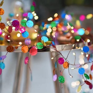 Puede incluir: Una guirnalda decorativa con lentejuelas redondas y coloridas en tonos rosa, naranja, verde, azul y morado. Las lentejuelas están unidas a un fino alambre marrón con pequeñas luces blancas cálidas. El fondo está desenfocado.