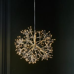 Puede incluir: Una lámpara decorativa esférica con numerosas luces LED pequeñas de color blanco cálido. La lámpara está suspendida de un fino cable blanco sobre un fondo oscuro. Las luces están dispuestas para parecerse a una explosión o una estrella.