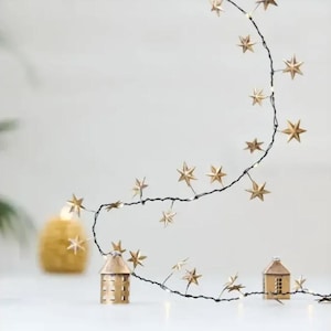 May include: A string of decorative lights featuring small, metallic gold-colored stars attached to a thin dark wire. The wire is draped across two small, house-shaped metal lanterns with cut-out windows, creating a warm and festive illumination.