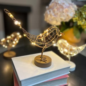 Puede incluir: Tres esculturas de pájaros de alambre dorado con luces LED blanco cálido. Los pájaros están sobre un libro blanco y una superficie marrón.