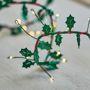 May include: A Christmas garland with green fabric holly leaves and small, warm white LED lights. The wire is wrapped with green and red twine, creating a festive holiday decoration.