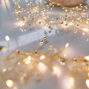 Peut inclure: Une guirlande lumineuse décorative composée de fines branches en fil métallique doré, ornées de petites perles sphériques transparentes. La structure en fil supporte de multiples minuscules ampoules LED aux tons chauds diffusant une lumière douce.
