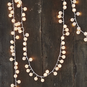 Pode incluir: Fio de luzes com lâmpadas esféricas brancas em um fio branco, contra um fundo de madeira escura. As luzes estão acesas, criando um brilho quente. Ideal para decoração festiva ou iluminação ambiente.