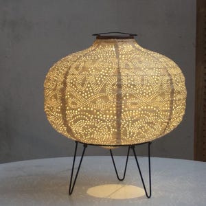 Op de afbeelding: Een witte keramische lamp met een delicaat patroon en een zwarte metalen standaard. De lamp is verlicht en geeft een warm licht.