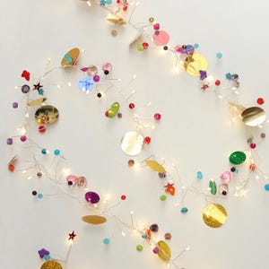 Puede incluir: Una guirnalda de luces decorativa con cuentas de colores, lentejuelas y detalles en forma de estrella. Las luces están ensartadas en un alambre fino y emiten un brillo cálido. La guirnalda presenta elementos dorados, rosas, rojos, verdes y azules.