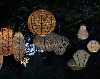 May include: Several decorative outdoor lanterns with intricate cut-out patterns hang from tree branches. The lanterns vary in shape, including round, oval, and geometric designs. The lanterns are lit, emitting a warm, inviting glow.