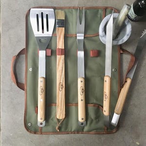 May include: A set of stainless steel grilling tools with light wood handles, including a slotted spatula, a basting brush, a two-pronged fork, a knife, and tongs, all organised in a green canvas roll-up case with brown trim and metal snaps.