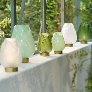 May include: A row of decorative glass candle holders in various shades of green and white. Each holder has a unique, textured design and sits on a gold-colored base. The holders are arranged on a white ledge with a blurred green background.