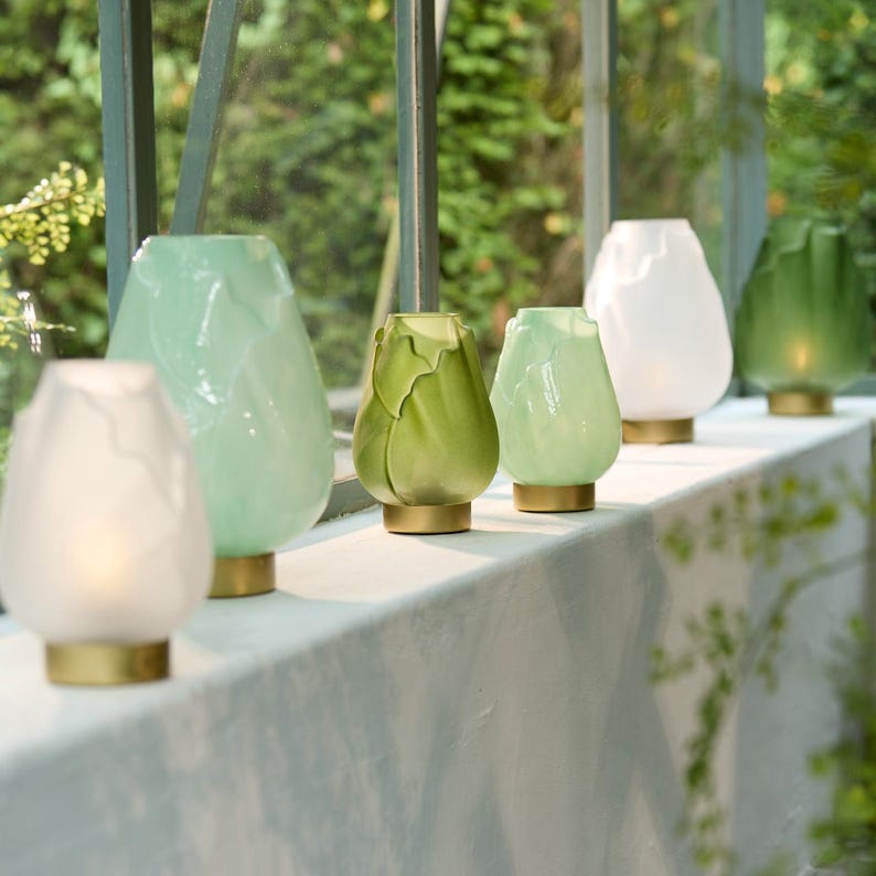 May include: A row of decorative glass candle holders in various shades of green and white. Each holder has a unique, textured design and sits on a gold-colored base. The holders are arranged on a white ledge with a blurred green background.