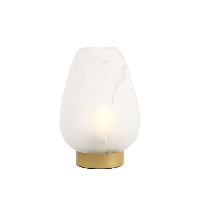 May include: A white, teardrop-shaped table lamp with a gold-colored base. The lamp has a frosted glass shade with a marbled pattern. The lamp is lit from within, creating a soft glow. The lamp is a decorative lighting fixture.