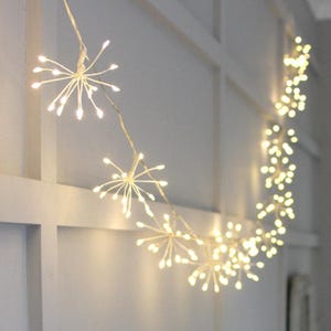 Puede incluir: Una cadena de luces blancas de hadas con un diseño de fuegos artificiales. Las luces están enhebradas en forma de curva y están encendidas, creando un brillo cálido.