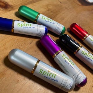 May include: An assortment of travel inhalers in various colors, including blue, green, red, black, purple, and silver. Each inhaler has a gold band and the words "Spirit travel inhaler" printed on a white label. The inhalers are arranged on a wooden surface.