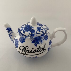 Puede incluir: Tetera de cerámica blanca con estampado floral azul y la palabra "Bristol" en escritura negra. La tetera tiene un asa curva, un pico y una tapa con un pomo. El diseño floral cubre el cuerpo y la tapa.
