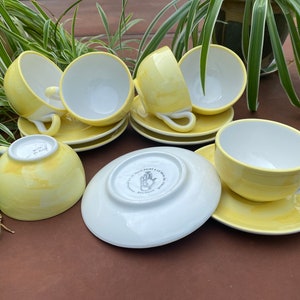 May include: Set of six yellow teacups and saucers with white interiors. The cups and saucers have a subtle marbled pattern. The saucers have a white underside with a black handprint logo.