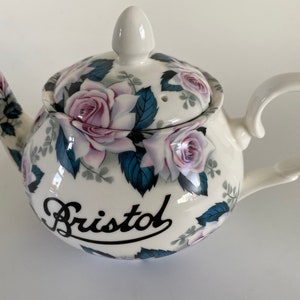 Pu&ograve; includere: Teiera in ceramica bianca con un design floreale con rose rosa e foglie blu. La teiera ha un coperchio e un manico. La parola "Bristol" &egrave; scritta in corsivo nero sul lato della teiera.