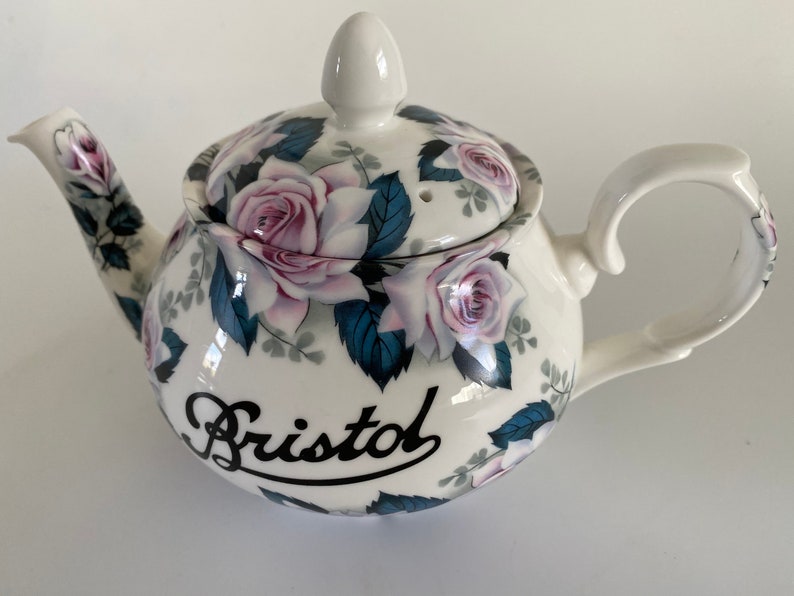 Pu&ograve; includere: Teiera in ceramica bianca con un design floreale con rose rosa e foglie blu. La teiera ha un coperchio e un manico. La parola "Bristol" &egrave; scritta in corsivo nero sul lato della teiera.