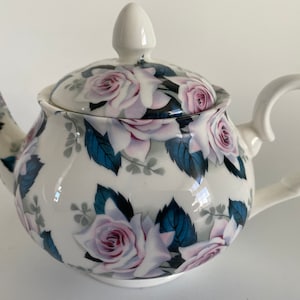 Puede incluir: Tetera blanca con un diseño floral con rosas rosas y hojas azules. La tetera tiene una tapa y un asa.