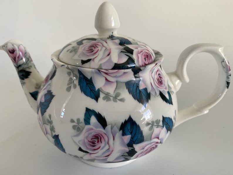 Pu&ograve; includere: Teiera bianca con un design floreale con rose rosa e foglie blu. La teiera ha un coperchio e un manico.