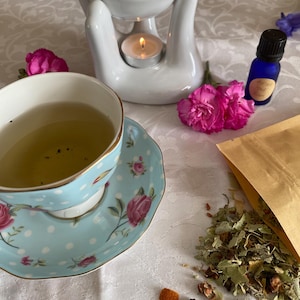 May include: A serene still life featuring a light blue teacup and saucer with a floral design, a white ceramic oil diffuser with a lit candle, and a small blue essential oil bottle. Pink carnations and loose tea leaves complete the scene, evoking a sense of calm.
