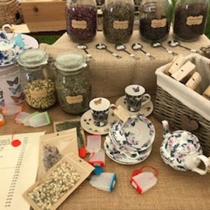 May include: A table display of loose leaf tea, tea bags, teacups, and a teapot. The teacups and teapot have a floral pattern with butterflies. The tea is in glass jars with labels. There are also tea infusers and a basket of soap.