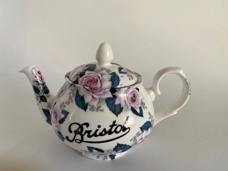 Pu&ograve; includere: Teiera bianca con un motivo floreale di rose rosa e foglie verdi. La teiera ha un'iscrizione nera che recita "Bristol".