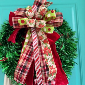 May include: A festive Christmas wreath with a green tinsel base. The wreath is adorned with a large bow featuring red, green, and white patterned ribbons, including plaid, polka dots, and poinsettia designs. A red velvet ribbon adds a touch of elegance.