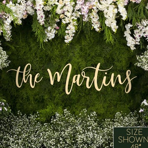 May include: A wooden sign with the text "the Martins" in gold script lettering. The sign is displayed against a backdrop of green foliage and white flowers. The sign is 48 inches wide.