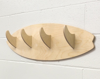 May include: A wooden surfboard-shaped wall mount with five fin-shaped hooks. The mount is made of light wood and is mounted on a white wall.