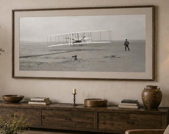 Könnte beinhalten: Ein gerahmtes Schwarz-Weiß-Foto des Wright Flyers im Flug, montiert an einer beigen Wand über einem langen, dunklen Medienboard aus Holz. Das Bild zeigt das historische Flugzeug über einer flachen Landschaft mit einer Person in der Nähe.