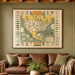 Op de afbeelding: Een grote rechthoekige wandprint getiteld Big Game Fish met een gedetailleerde kaart van Noord-Amerika omringd door illustraties van diverse vissoorten, ingelijst in een dunne houten rand en opgehangen aan een olijfgroene muur.