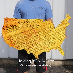 Puede incluir: Un recorte de metal amarillo de los Estados Unidos de América con el texto "We the People" y el preámbulo de la Constitución impreso en él. El recorte está siendo sostenido por una persona. El texto debajo del recorte dice "Holding 35" x 24" size (Smaller sizes available)"