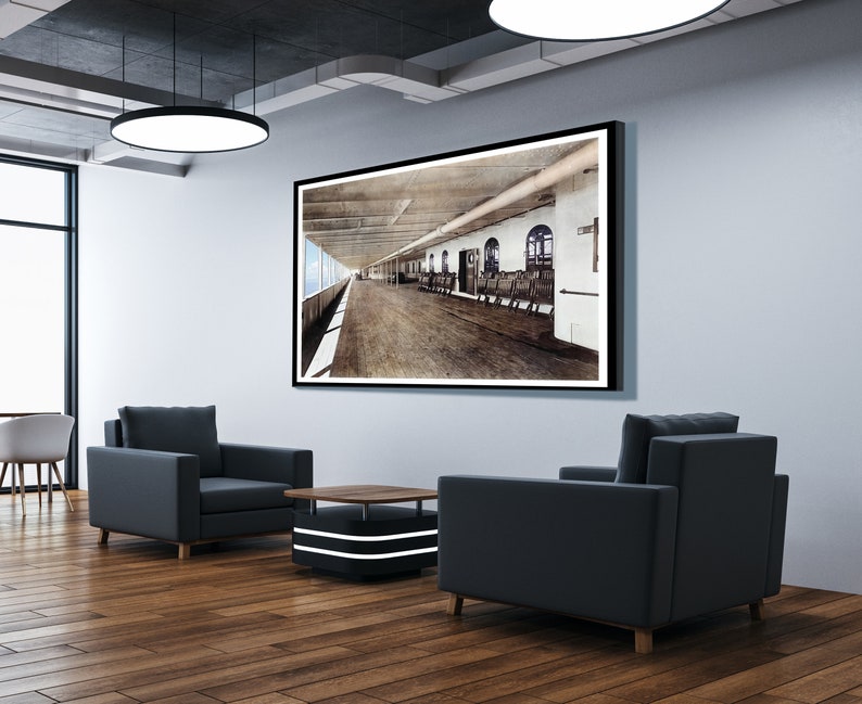 Op de afbeelding: Een zwart-witfoto van een scheepsdek, ingelijst en hangend aan een muur in een moderne kantooromgeving. Twee zwarte fauteuils en een salontafel staan voor het beeld.