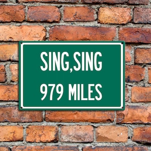 May include: Green road sign with white text that reads "Sing Sing 979 Miles". The sign is mounted on a brick wall.