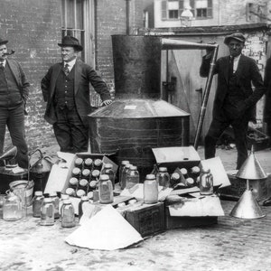 May include: A black and white photo of four men standing in front of a large metal container. The men are wearing suits and hats. The container is surrounded by various glass jars and metal funnels. The scene appears to be a still from a historical photo of a moonshine operation.