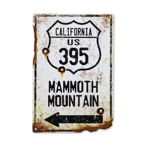 May include: A white metal sign with a black arrow pointing left. The sign has a vintage look with rust and wear. It reads "California US 395 Mammoth Mountain".