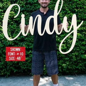 May include: A person holding a large, white wooden sign with the name "Cindy" written in a cursive font. The sign is 48 inches tall and is shown in font #10.