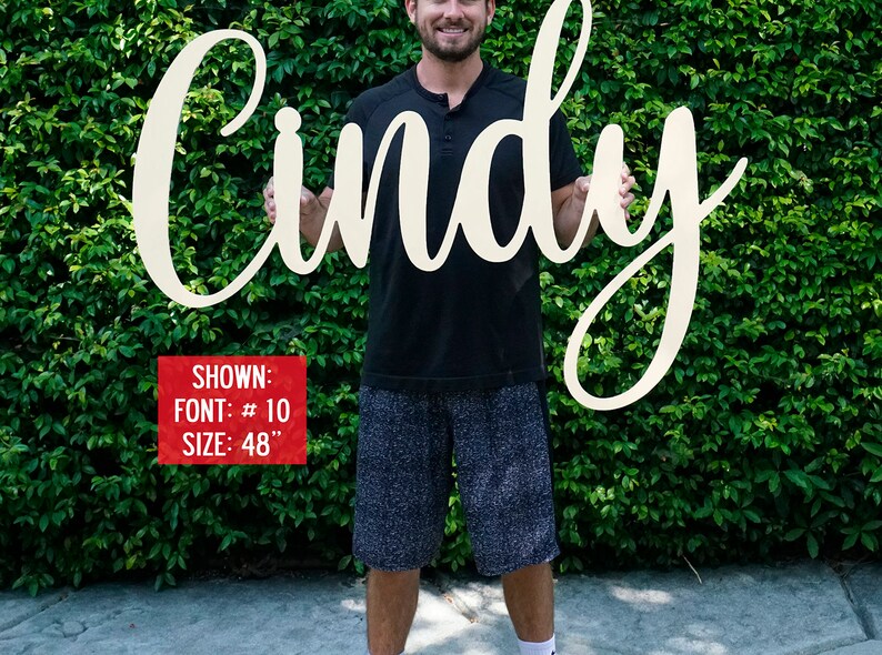 May include: A person holding a large, white wooden sign with the name "Cindy" written in a cursive font. The sign is 48 inches tall and is shown in font #10.