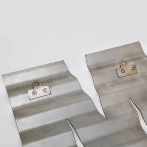 May include: Two corrugated metal pieces with mounting brackets. The metal has a brushed finish and a wavy pattern. The brackets are rectangular with two screw holes and a single circular hole. The background is plain white.