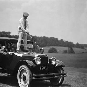 May include: A black and white photo of two men playing golf. One man is standing on the hood of a vintage car, swinging a golf club. The other man is standing on the ground, watching the golfer. The car has a license plate that reads "D-6-567".