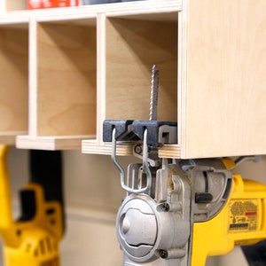 May include: A yellow and black jigsaw with a silver blade is hanging from a wooden shelf. The jigsaw is attached to the shelf with a black and silver hook.